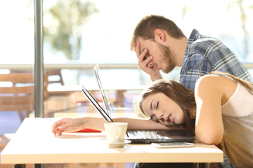 A tired woman resting her head on a desk, surrounded by a laptop and coffee, representing fatigue due to lifestyle factors.